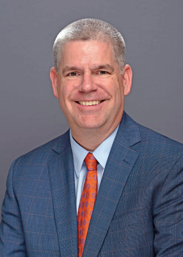 Dr. William “Bill” Fiege in a dark blue suit jacket, light blue shirt,  and an orange tie in front of a blue background.