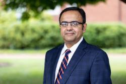 The image is of a man with short black hair, wearing glasses, a dark blue suit, a white dress shirt, and a striped tie in shades of orange and blue. He is standing outdoors with a blurred background of greenery. The expression on his face is neutral, and he is looking directly at the camera.