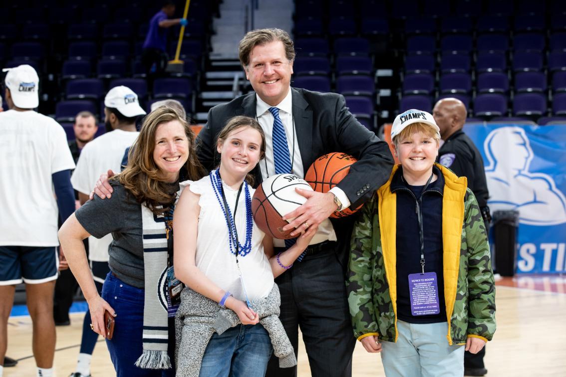 Reveley Family at the Big South Tournament - Group of four people posing together on a basketball court. A man in a suit holds basketballs while standing with a woman and two children. They are smiling at the camera in an arena with seating in the background. Event credentials, team gear, and championship hats suggest a post-game celebration.