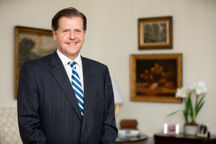 W. Taylor Reveley IV - Portrait of a man wearing a dark pinstripe suit and striped tie. He is smiling and facing the camera in an indoor office setting. His hair is neatly styled and combed back. Framed artwork and a plant appear softly blurred in the background.