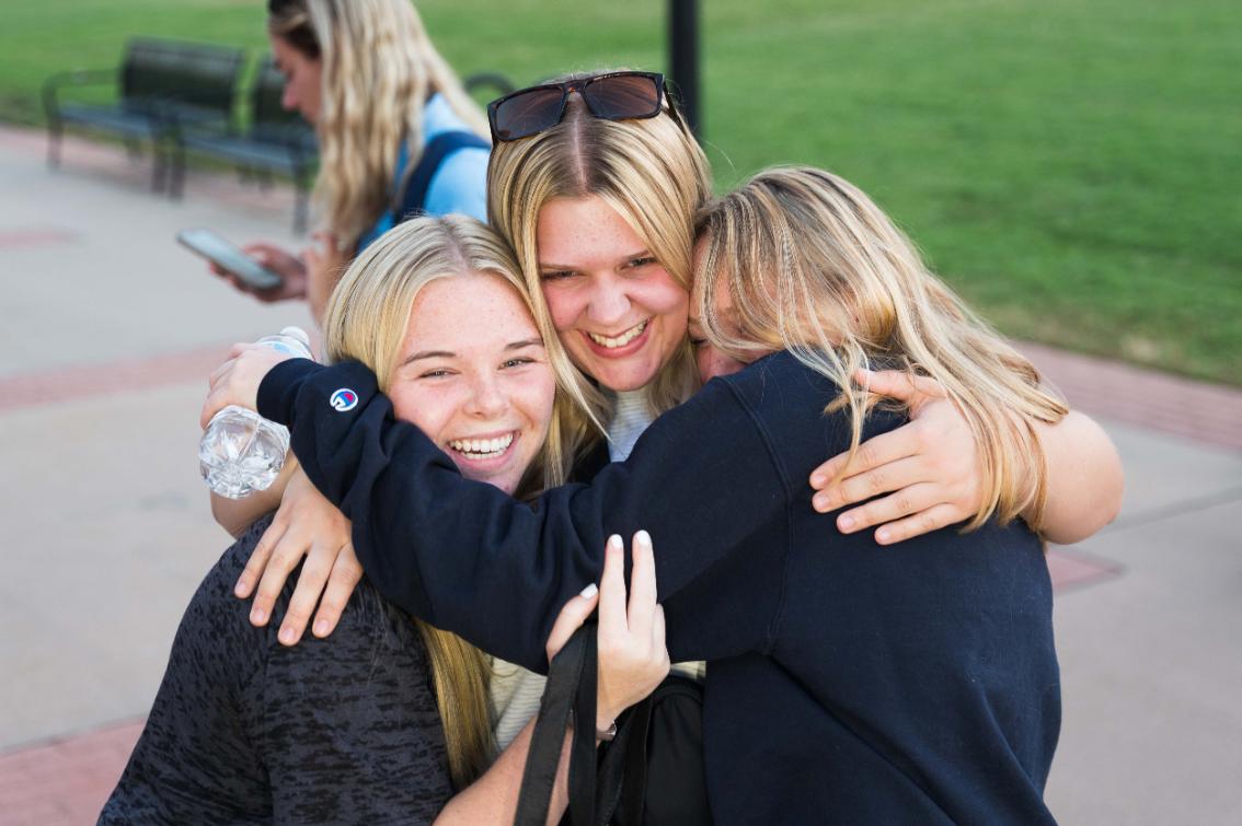 Three students hugging during New Lancer Days - Three students hug closely together and smile on a campus walkway. One student holds a water bottle while the others wrap their arms around her. A lawn and benches appear in the background. Friends share a joyful moment during a campus event.