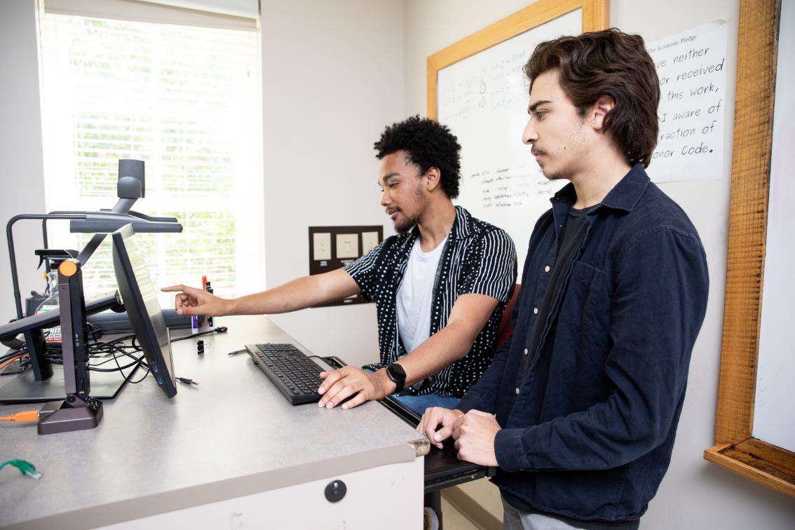 Two students working together on a project - Two students work together at a computer station in a classroom. One student points at the monitor while explaining something. A keyboard, document camera, and whiteboard are visible nearby. Students collaborate on coursework using classroom technology.