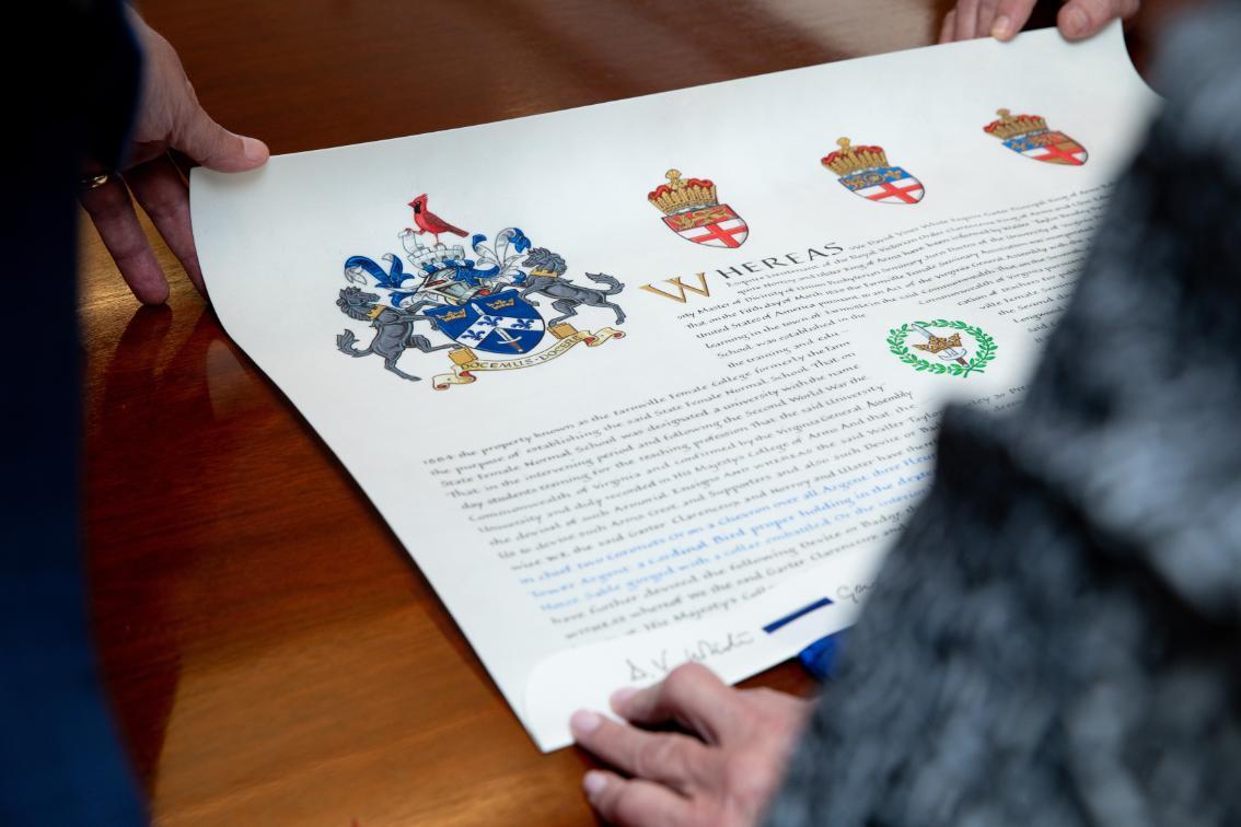 Longwood’s new coat of arms is displayed on a formal document as people review it at a table.