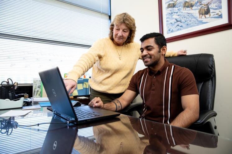Two people collaborate at a desk while looking at a laptop in an office. A woman in a yellow sweater stands beside a seated man and points toward the screen. The man smiles while using the laptop at a glass-topped desk. Office items, window blinds, and a framed wildlife print hang on the wall behind them.